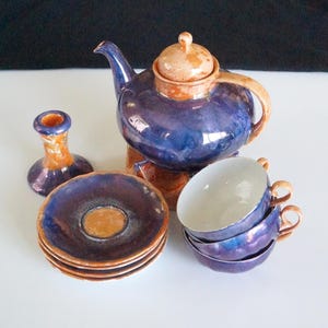 May include: A ceramic tea set with a blue and orange glaze. The set includes a teapot, four saucers, four teacups, and a small candle holder. The teapot has a lid and a curved handle. The teacups are stacked. The saucers are stacked.