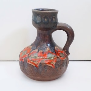 Pode incluir: Um jarro de cerâmica castanho com uma pega. O jarro apresenta um topo castanho escuro com uma série de círculos em relevo. O corpo do jarro tem um design floral vermelho e verde. A base do jarro tem um padrão em relevo.