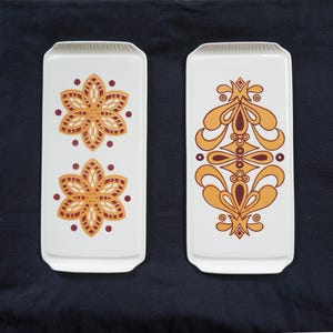 Könnte beinhalten: Zwei rechteckige Serviertabletts aus weißer Keramik mit dekorativen Mustern. Ein Tablett zeigt zwei stilisierte Blumenmuster in Orange- und Brauntönen. Das andere Tablett zeigt ein stilisiertes geometrisches Muster in Orange- und Brauntönen.