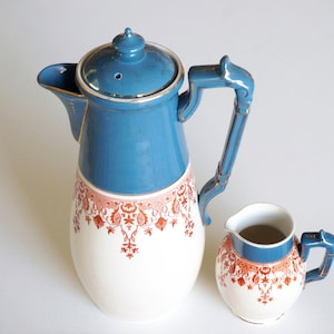 Könnte beinhalten: Ein blaues und weißes Keramik-Kaffee- und Milchkännchen-Set mit floralem Design. Die Kaffeekanne hat einen Deckel und einen Griff mit goldenen Akzenten. Der Milchkännchen hat einen Griff und einen blauen Rand.