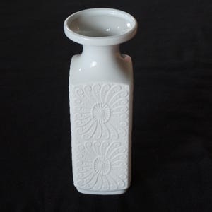 May include: White ceramic vase with an embossed floral design. The vase has a square body and a narrow neck.