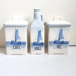 Op de afbeelding: Set van drie keramische keukencontainers en een fles met een nautisch thema. De witte containers hebben blauwe zeilbootillustraties en de woorden "CAFE", "HUILE" en "RIZ". De fles heeft een handvat en een bijpassend zeilbootontwerp.