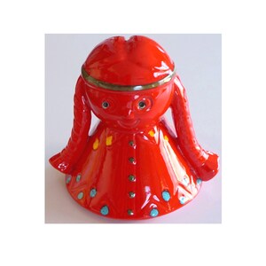 Può includere: Salvadanaio in ceramica rossa a forma di ragazza con trecce. La ragazza ha un viso sorridente con occhi blu e accenti gialli. Il salvadanaio è decorato con punti blu e turchesi.