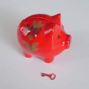 Op de afbeelding: Een glanzend rood spaarvarken met een gleuf aan de bovenkant en een kleine sleutel. Het spaarvarken heeft een bloemmotief in bruin- en goudtinten. De sleutel is rood. Het spaarvarken is een klassiek item om geld te sparen.