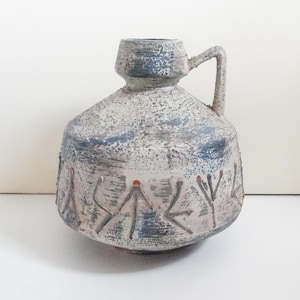 Puede incluir: Un jarrón de cerámica con asa, con una superficie texturizada y moteada en tonos grises, azules y rosas. El jarrón tiene un cuerpo bulboso con cuello cilíndrico y está decorado con símbolos estilizados y pequeños acentos rojos.