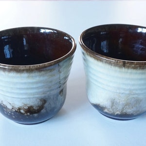 May include: Two ceramic cups with a distinctive design. The cups feature a dark brown interior and a white exterior with brown accents. The cups have a slightly angled rim and a smooth, glossy finish. These cups are suitable for serving drinks.