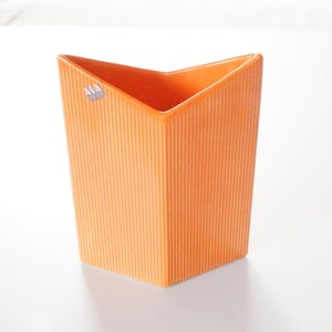 Op de afbeelding: Een oranje, rechthoekige vaas met een textuur, verticaal geribbeld ontwerp. De vaas heeft een hoekige bovenkant en een klein label in de linkerbovenhoek. De vaas is waarschijnlijk van keramiek of een vergelijkbaar materiaal.
