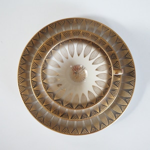May include: Overhead view of a white teacup and saucer set with gold floral and dot patterns. The cup has a small handle and sits on a matching saucer and a larger plate, all with a gold rim.