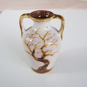 以下が含まれることがあります： 2つの取っ手が付いた小さな白い陶器の花瓶。花瓶は茶色の釉薬がかけられており、茶色の枝とピンクの花が咲いた木の絵が描かれています。