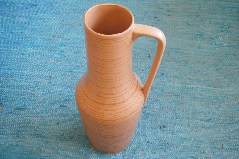 Puede incluir: Un jarr&oacute;n de cer&aacute;mica marr&oacute;n claro, alto, con una boca ancha y un asa. El jarr&oacute;n tiene un patr&oacute;n de veta de madera y est&aacute; sobre una superficie azul.