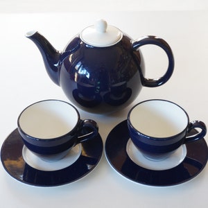 Op de afbeelding: Een donkerblauwe theepot met een wit deksel en twee bijpassende theekopjes en schoteltjes. De theepot en kopjes hebben een witte binnenkant.