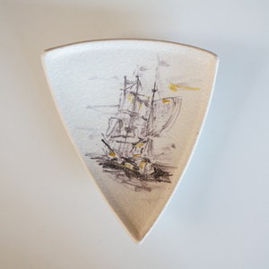 May include: A white ceramic triangular dish with a black and gold illustration of a sailboat on the front.