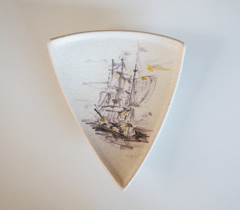 May include: A white ceramic triangular dish with a black and gold illustration of a sailboat on the front.