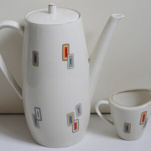 May include: A white ceramic coffee pot and creamer set with a geometric pattern in shades of blue, green, yellow, and orange. The pot has a lid and a handle. The creamer has a handle.