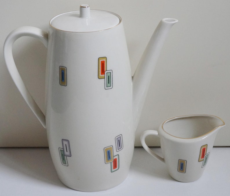 May include: A white ceramic coffee pot and creamer set with a geometric pattern in shades of blue, green, yellow, and orange. The pot has a lid and a handle. The creamer has a handle.