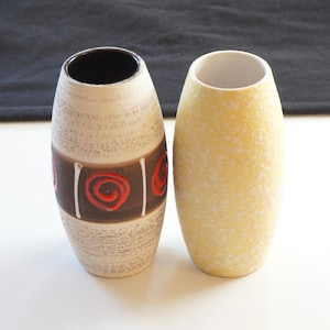 Op de afbeelding: Twee keramische vazen, een met een wit en bruin gespikkeld glazuur en rode en witte swirl-ontwerpen, en de andere met een geel gespikkeld glazuur.