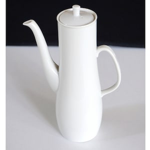 Op de afbeelding: Een witte koffiekan van keramiek met een deksel en een handvat. De kan heeft een hoge, slanke vorm.