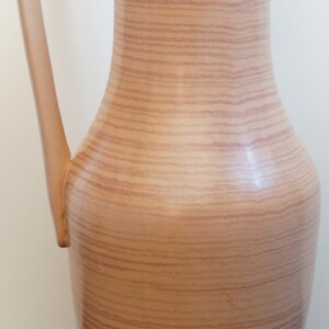 Puede incluir: Un jarr&oacute;n alto de cer&aacute;mica marr&oacute;n con un patr&oacute;n de veta de madera. El jarr&oacute;n tiene una base ancha y un cuello estrecho con un asa curva.