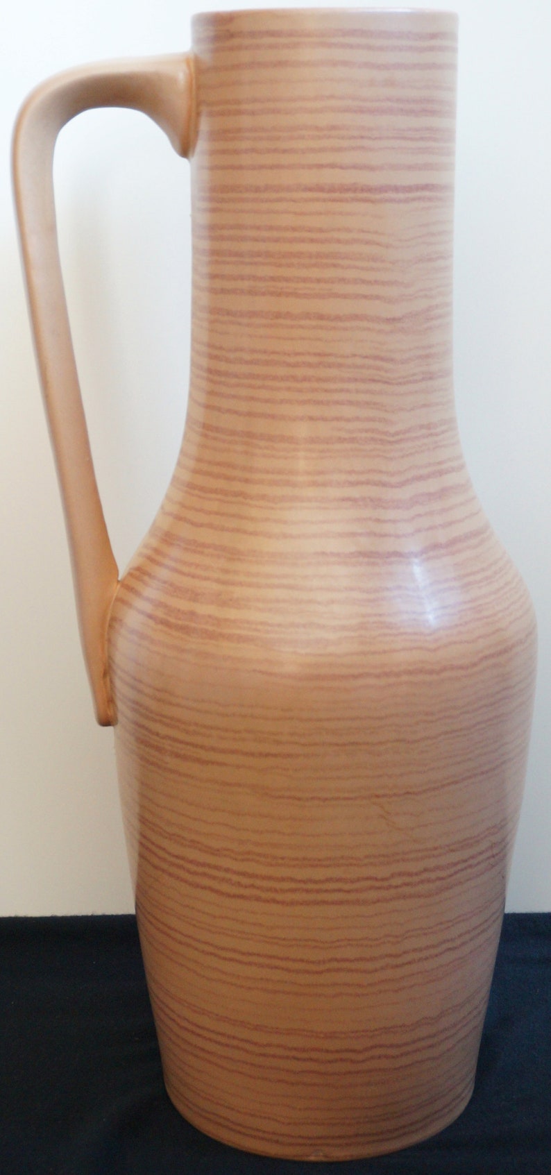 Puede incluir: Un jarr&oacute;n alto de cer&aacute;mica marr&oacute;n con un patr&oacute;n de veta de madera. El jarr&oacute;n tiene una base ancha y un cuello estrecho con un asa curva.