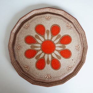May include: A round ceramic plate with a brown and white speckled glaze. The centre of the plate features a stylised flower design with orange petals and a brown centre.