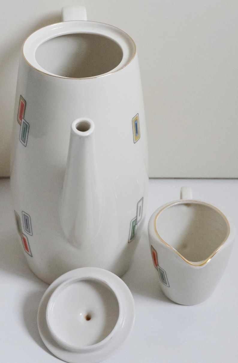 May include: A white ceramic coffee pot with a lid and a matching creamer. The pot has a geometric pattern of colorful squares.