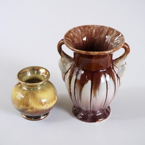 Könnte beinhalten: Zwei Keramikvasen, eine mit brauner und weißer Glasur und die andere mit gelber und brauner Glasur. Die größere Vase hat zwei Henkel und ein geriffeltes Design.