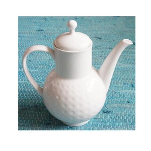 Op de afbeelding: Een witte keramische theepot met een deksel en een gebogen handvat. De body van de theepot heeft een getextureerd, kuiltjespatroon. De theepot staat tegen een blauwe, getextureerde achtergrond.