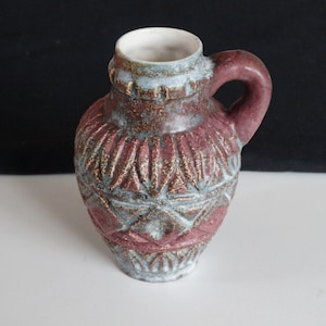 Può includere: Un piccolo vaso in ceramica marrone e bianco con un motivo geometrico. Il vaso ha un manico e un corpo arrotondato.