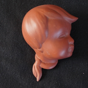Op de afbeelding: Een terracottakleurig keramisch beeld van een kinderhoofd met een enkele vlecht. Het beeld is een zijaanzicht van het gezicht van het kind, met gesloten ogen en een licht geopende mond.