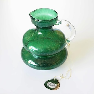 May include: A green glass pitcher with a clear handle and spout. The pitcher has a textured surface and a round base. A small tag is attached to the pitcher with a string.