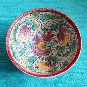Könnte beinhalten: Eine weiße Keramikschüssel mit rotem Rand und einem farbenfrohen Blumendesign. Die Schüssel zeigt einen stilisierten Vogel mit grünen, roten, gelben und blauen Akzenten. Das Design erinnert an traditionelle italienische Majolika-Keramik.