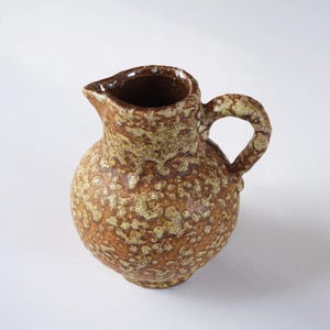 以下が含まれることがあります： 斑点のある釉薬をかけた小さな茶色の陶器のピッチャー。ピッチャーは丸いボディとハンドルを持っています。