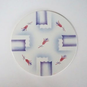 May include: A round, white decorative plate with a repeating pattern. The design features purple and blue geometric shapes and red floral accents. The plate is a decorative item.