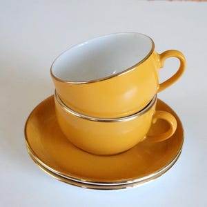 Op de afbeelding: Twee gestapelde mosterdgele keramische theekopjes met witte binnenkant en goudkleurige randen, rustend op twee bijpassende mosterdgele schoteltjes met goudkleurige randen. De kopjes hebben gebogen oren en een gladde, effen gekleurde afwerking.