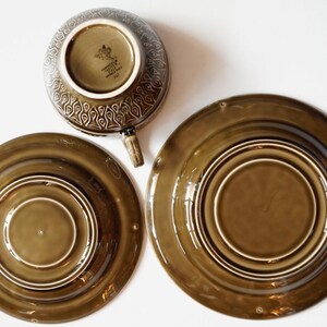 May include: A set of three brown ceramic plates with a concentric circle design. The largest plate is 25 cm in diameter. The smallest plate is 15 cm in diameter. The cup is 10 cm in diameter. The cup has a brown glaze with a floral design and the text "Made in England" on the bottom.