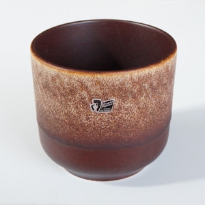 May include: A brown ceramic cup with a textured, speckled exterior and a smooth, dark brown interior. The cup has a slightly flared rim and a small sticker on the side. The cup is likely for drinking beverages.
