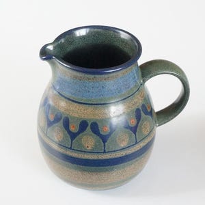 Op de afbeelding: Een keramische kan met een gespikkelde blauwe en beige glazuurlaag, voorzien van een donkerblauwe rand en handvat. Het lichaam is versierd met een herhalend patroon van donkerblauwe gestileerde bloemvormen en kleine oranje stippen in een band.
