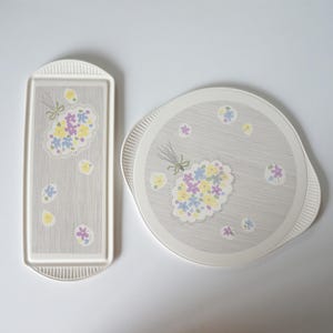 Op de afbeelding: Twee vintage serveerschalen met een bloemenmotief. De grotere schaal is rond en de kleinere is rechthoekig. Beide schalen hebben een grijze achtergrond met een witte rand en een bloemenmotief in tinten geel, blauw en paars.