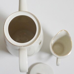 May include: A white porcelain teapot with a gold trim and a matching creamer and lid. The teapot has a spout and a handle. The creamer has a handle and a spout. The lid has a knob.