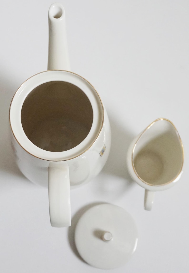 May include: A white porcelain teapot with a gold trim and a matching creamer and lid. The teapot has a spout and a handle. The creamer has a handle and a spout. The lid has a knob.