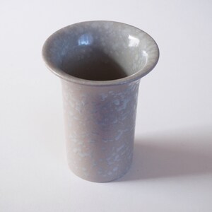 May include: A light grey ceramic vase with a speckled glaze. The vase is cylindrical in shape and has a slightly flared opening.