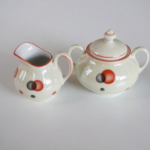 May include: A cream-coloured ceramic creamer and sugar bowl set. The set features red and black polka dots and red trim. The sugar bowl has a lid with a small knob. The creamer has a handle and a spout.