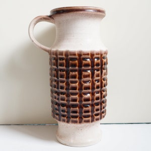 Könnte beinhalten: Eine weiße Keramikvase mit einem braunen geometrischen Muster. Die Vase hat einen Henkel und einen abgerundeten Boden.