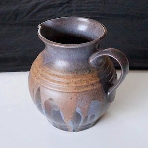 Può includere: Brocca in ceramica fatta a mano con corpo arrotondato e manico. La brocca presenta una smaltatura maculata nei toni del marrone, blu e beige. Ha un beccuccio per versare ed è adatta per servire bevande. Oggetto decorativo.