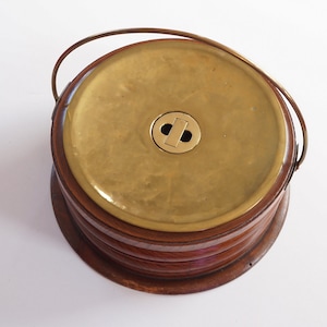 Op de afbeelding: Een vintage ijsemmer met een messing deksel en handvat, en een houten body. De emmer heeft een cirkelvorm met een messing sluiting op het deksel. De houten body heeft een gelaagd ontwerp. De handgreep is van messing.
