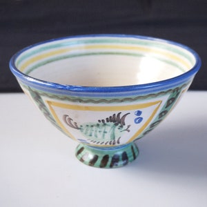 Op de afbeelding: Een kleine, witte keramische kom met een blauwe rand en een groen, geel en blauw visontwerp. De kom heeft een klein, groen en zwart voetje.