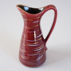 May include: A small red ceramic jug with a white and gold geometric pattern. The jug has a wide base and a narrow neck.