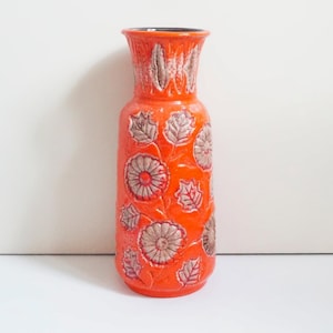 Op de afbeelding: Een hoge, oranje keramische vaas met een textuur en een bloemenreliëf. De vaas heeft een cilindrische vorm met een uitlopende rand en een donker interieur. Het reliëfpatroon omvat gestileerde bloemen en bladeren in een contrasterende beige kleur.