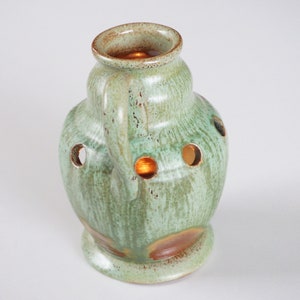 May include: A green ceramic vase with a brown glaze and a handle. The vase has holes around the sides and a rounded base.