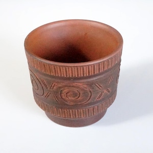 Op de afbeelding: Een terracotta-kleurige keramische pot met een brede opening en een decoratieve band met spiraal- en kruisachtige patronen. De pot heeft een licht uitlopende basis en een getextureerd oppervlak.